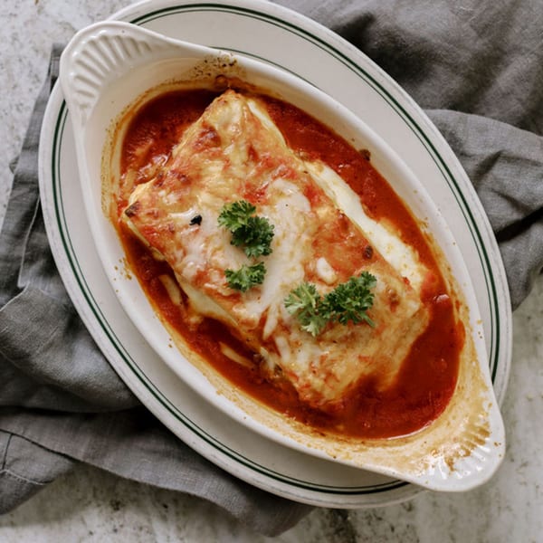 Baked lasagna with melted cheese and tomato sauce, garnished with parsley in a white dish.