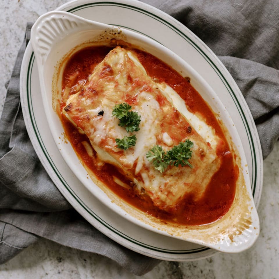 Baked lasagna with melted cheese and tomato sauce, garnished with parsley in a white dish.
