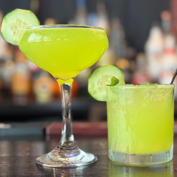 Two vibrant green cocktails, one in a coupe glass and one in a rocks glass, garnished with cucumber slices.