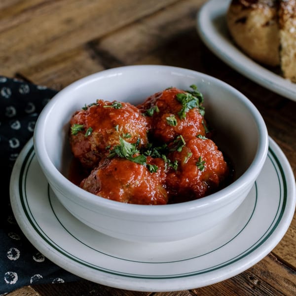 Three Frittella Italian Cafe meatballs in red sauce, garnished with parsley, in a white bowl.
