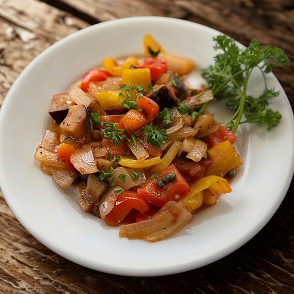 A white bowl filled with colorful Italian caponata, garnished with fresh parsley, on a wooden table.