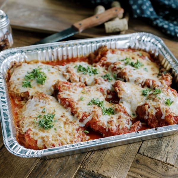 Chicken parmesan in a foil pan, topped with melted cheese, tomato sauce, and fresh parsley.